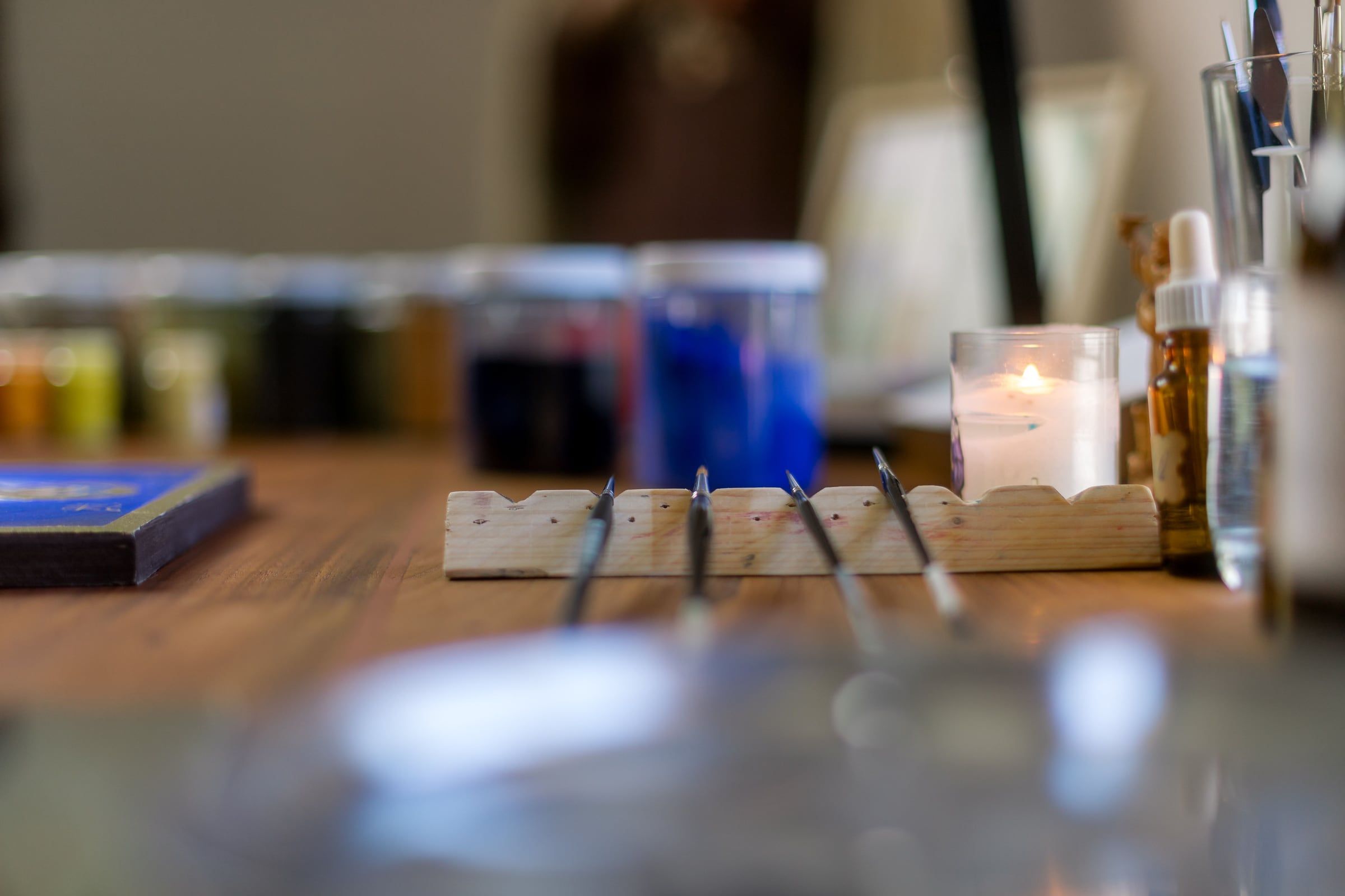 La table de travail de l’atelier : des pinceaux fins posés sur un repose-pinceaux en bois, des pots de pigments, une bougie allumée et le bord d’une icône